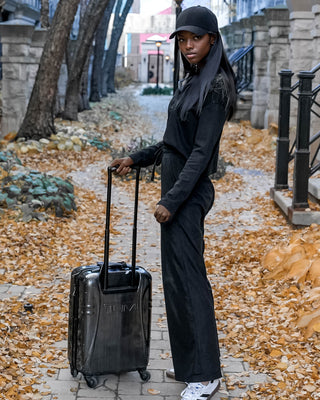 A slender black woman in a black hat, pullover, and pants, stands with a black roller suitcase amidst a picturesque fall city landscape, turned to her right but looking back at the camera over her left shoulder.