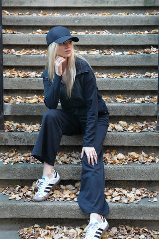 A blonde woman, dressed in a black hat, black pullover, and black pants paired with white and black gym shoes, sits on a concrete stoop scattered with fall leaves, gazing to her left, blending urban fashion with autumnal charm.