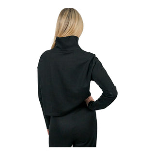 Viewed from the back, a woman with blonde highlights stands with one hand on her hip, wearing a black EMF protective turtleneck, zip-up its high collar and snug fit illustrating the garment's protective function against electromagnetic fields.