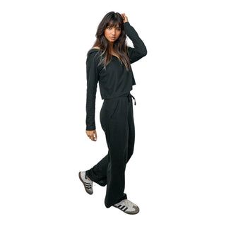 A woman with long, dark hair stands casually, one hand raised to her head, wearing a relaxed black EMF protective tracksuit paired with classic sneakers, highlighting the outfit's casual yet purposeful design for everyday electromagnetic field protection.