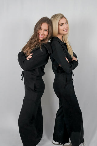 Two in girls stand back-to-back, smiling at the camera, wearing sleek black electromagnetic field (EMF) protective apparel; the clothing appears comfortable yet fashionable, designed to shield the body from electromagnetic radiation while maintaining a stylish look.